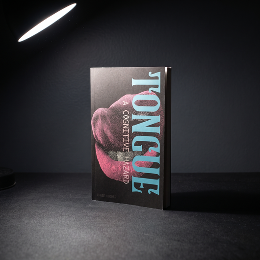 TONGUE: A Cognitive Hazard paperback standing upright on a black desk under a single lamp
